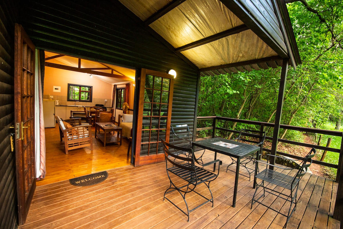 An inviting porch area features a wooden deck with a table and chairs, ideal for outdoor relaxation. The chalet interior is visible, showcasing a cozy living space with comfortable seating and natural light flowing through the large windows, surrounded by lush greenery.