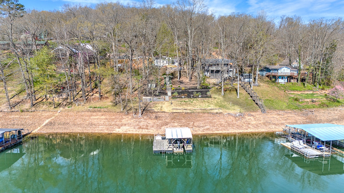 A serene view of the lakefront property reveals a gently sloping shoreline bordered by trees. Multiple decks and steps lead up from the water, where boat docks are seen anchored in the calm waters of Beaver Lake.