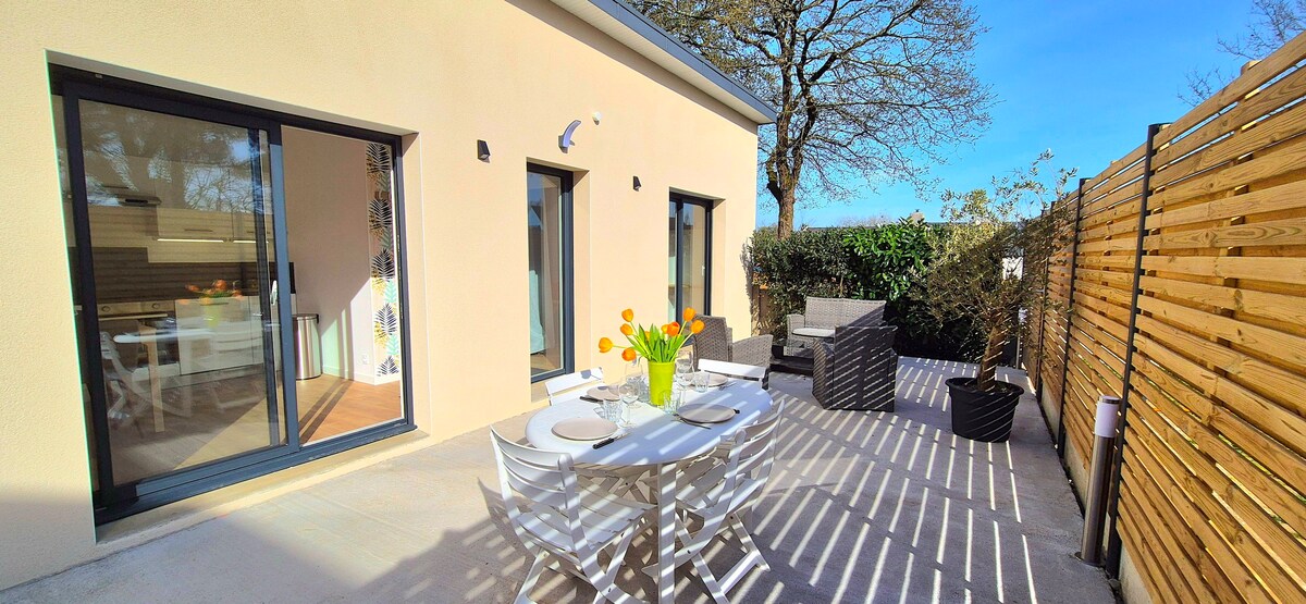 A patio area is shown, featuring a white dining table set with plates and a vase of fresh tulips. Comfortable seating is arranged nearby, with sunlight casting shadows from the wooden fencing and trees surrounding the space.