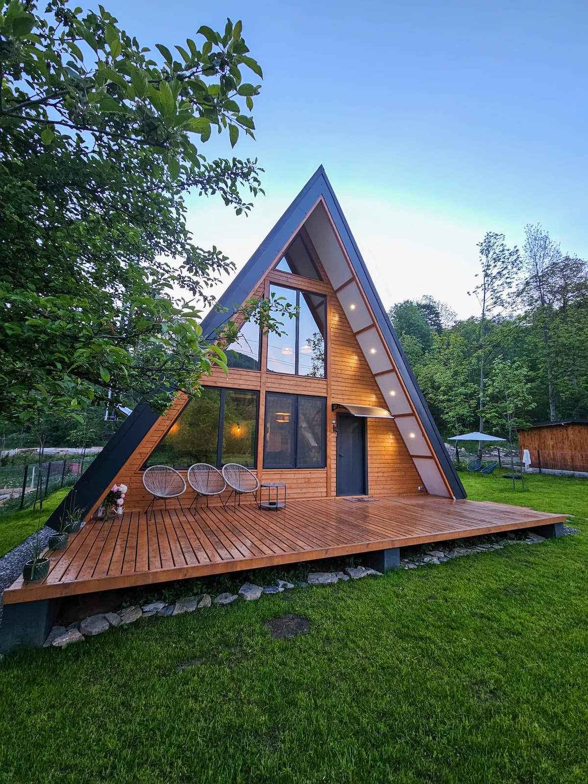 The A-frame chalet is surrounded by lush greenery, featuring large windows that provide natural light. A spacious wooden deck is seen at the entrance, with stylish seating arrangements and decorative elements, inviting guests to enjoy the serene surroundings.