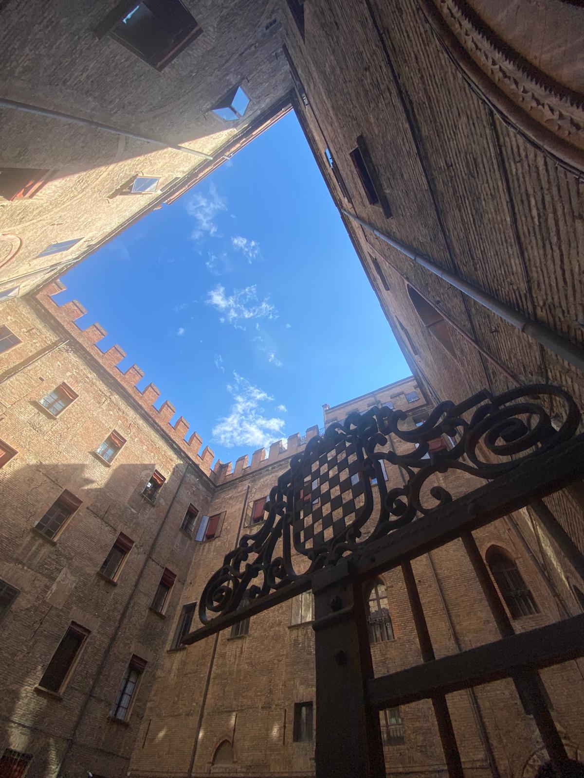 The inner courtyard of a historic building features a tall brick wall surrounding a clear blue sky, with a wrought iron gate in the foreground. The brickwork showcases intricate historical details, while the sunlight highlights the texture of the walls.
