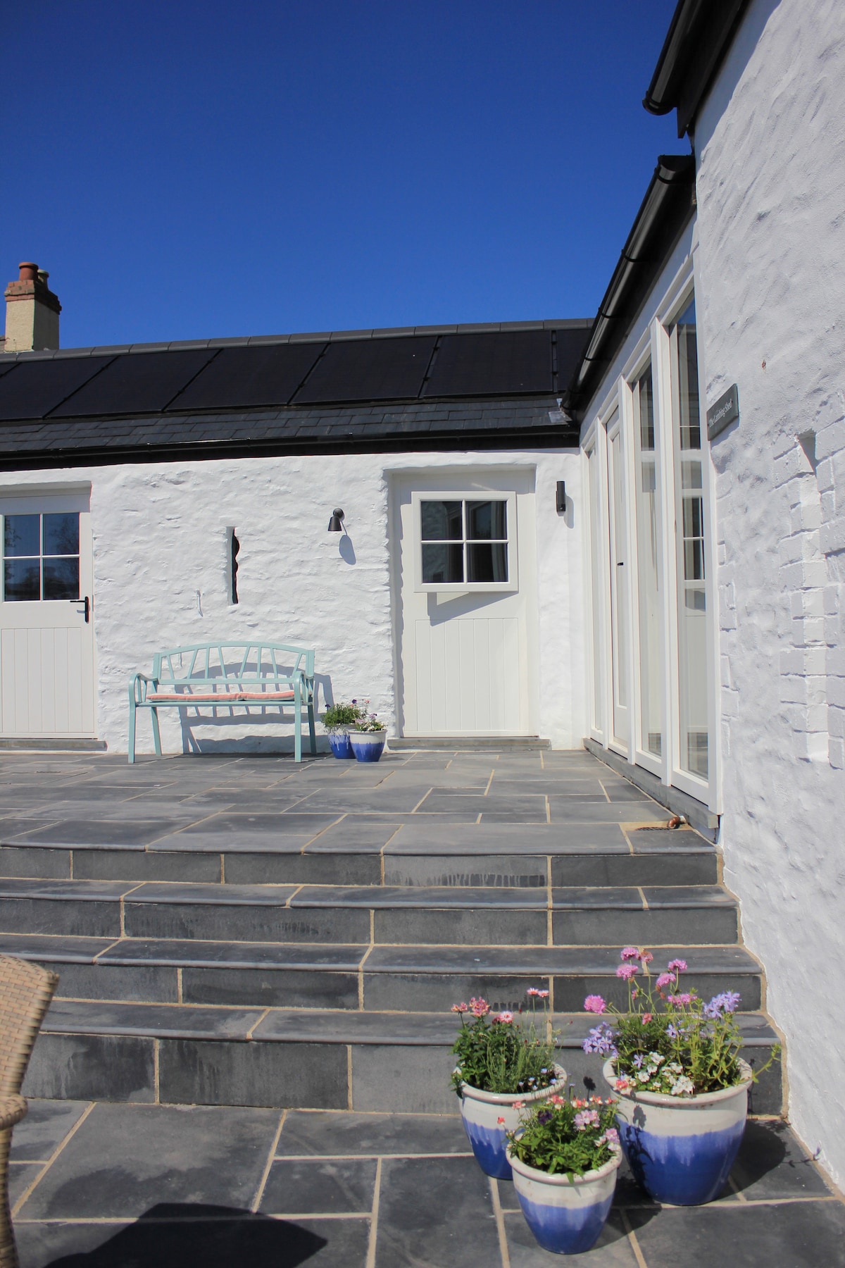 A sunlit patio area features stone steps leading up to the entrance of the cottage. Adorned with potted flowers, the space offers a serene atmosphere. A light blue bench is positioned against the whitewashed wall, which complements the clear blue sky above.