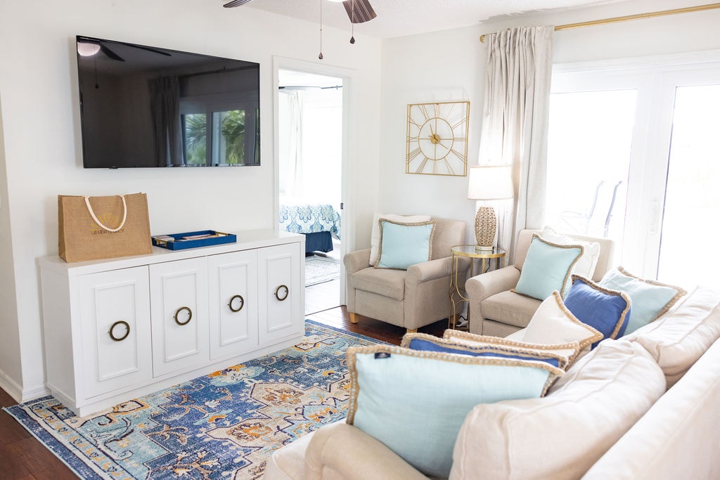 A modern living area is featured with comfortable seating arranged around a patterned rug. A large television is mounted on the wall, while natural light enters through sliding glass doors, providing a view of the outdoors. Decorative accents include a side table with a lamp.