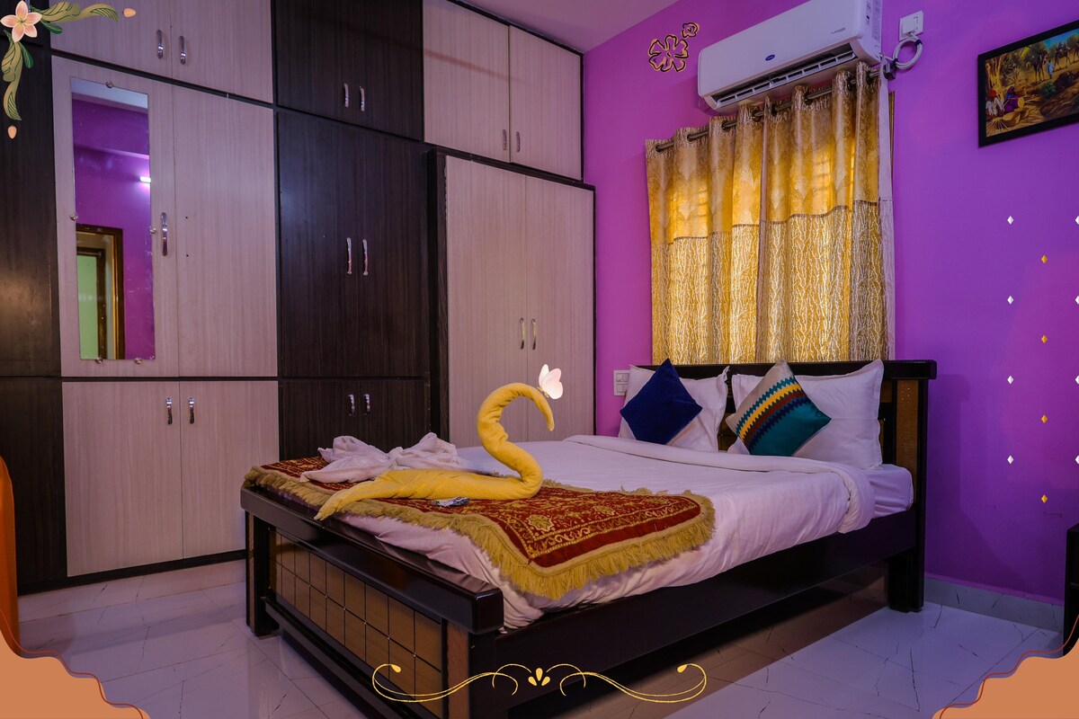 A cozy bedroom featuring a spacious bed with decorative pillows and a warm throw. Ambient lighting filters through the golden curtains, enhancing the vibrant purple walls. A large wardrobe offers ample storage, while a wall-mounted air conditioner ensures comfort.