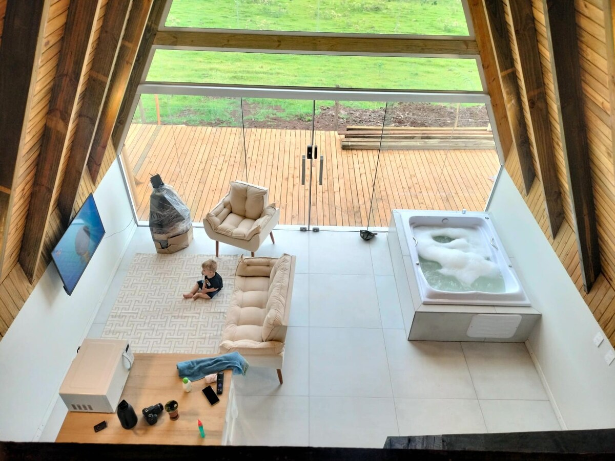 An aerial view of a spacious living area features a light-colored sofa and a large hot tub. A child plays on a textured rug, while glass doors provide a view of the outdoor deck. Natural light floods the space, highlighting the wood-beamed ceiling.