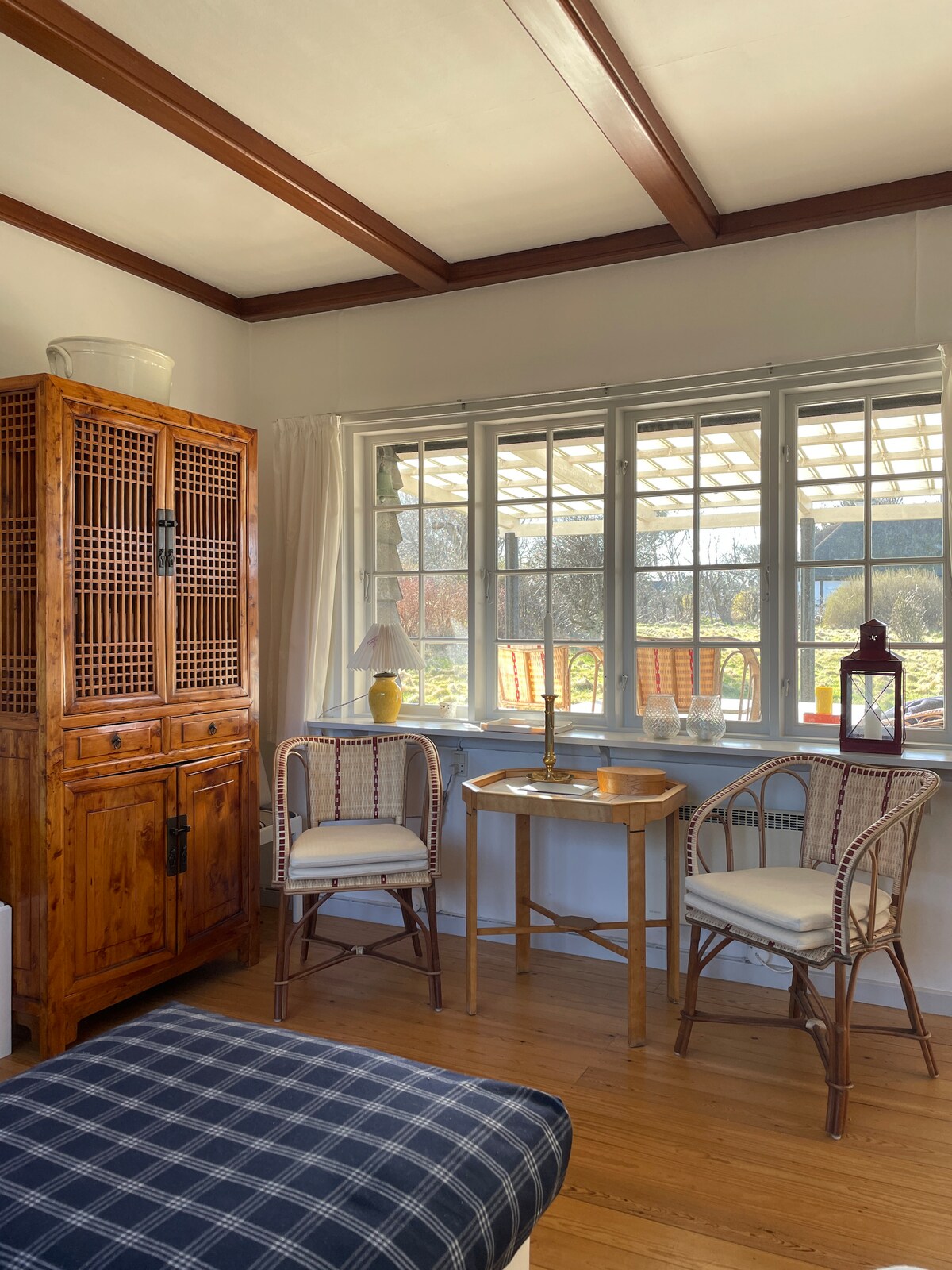 A cozy corner features a wooden cabinet paired with two rattan chairs and a small round table. Natural light filters through multiple windows, illuminating the space. Soft furnishings and decorative accents add a homey feel to the tranquil atmosphere.