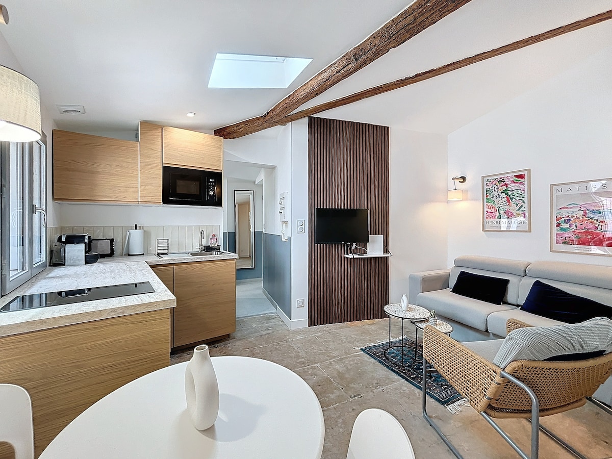 An open-plan kitchen and living space is highlighted by natural light from a skylight. Light wood cabinetry and sleek appliances complement the modern decor. A circular dining table is positioned alongside a cozy seating area, which features a minimalist sofa and decorative chair.