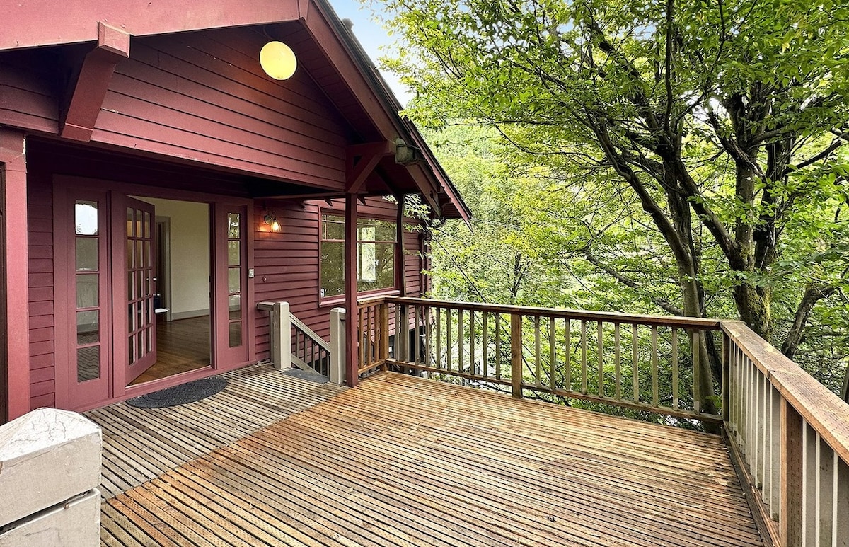 An outdoor deck with wooden flooring is shown, bordered by a railing. The entrance features double French doors, allowing access to the interior. Nearby trees provide natural foliage, creating a serene atmosphere. Soft natural light filters through the greenery.
