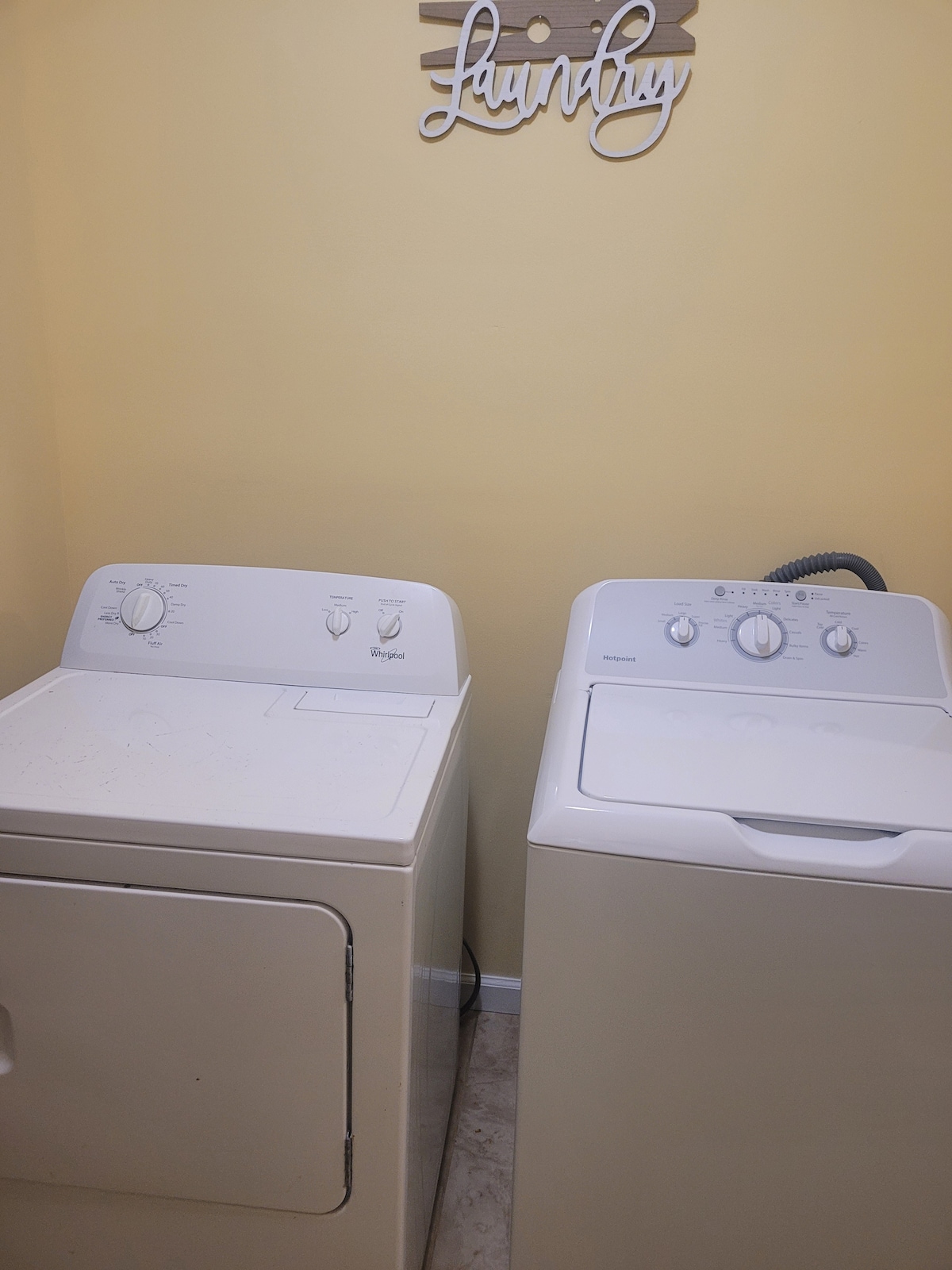 A dedicated laundry area is equipped with a washer and dryer side by side against a light yellow wall. A wooden sign with the word 'Laundry' is mounted above them, adding a functional yet homey touch to the space.