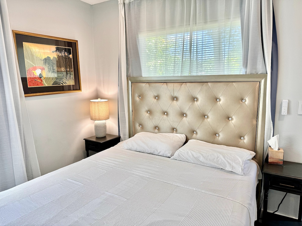A serene sleeping area is featured, showcasing a tasteful light-colored headboard with tufted detailing. Natural light filters through sheer curtains, illuminating the bed, which is dressed with crisp white linens. A bedside lamp and framed artwork add subtle elegance to the space.