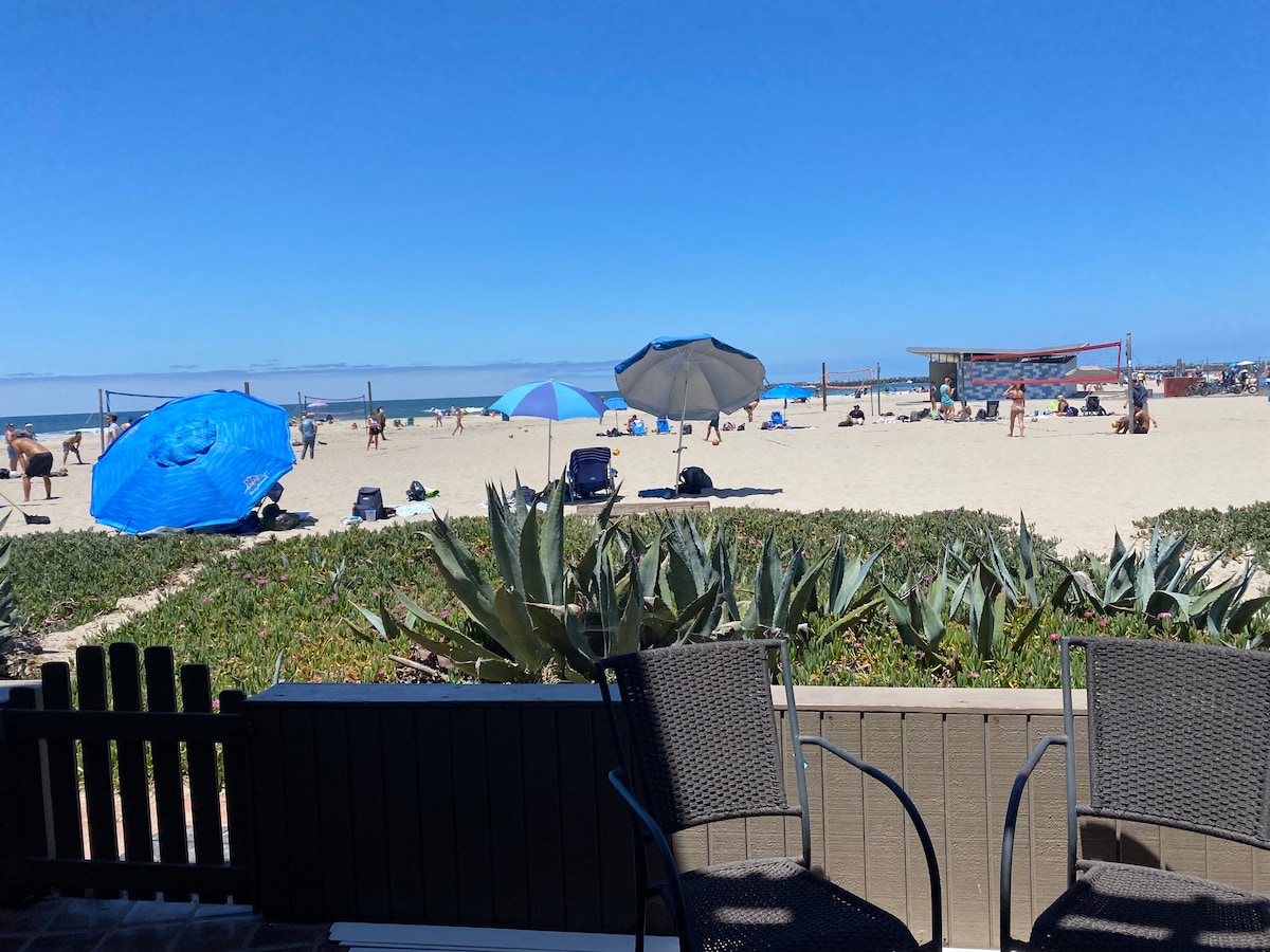 A view from the patio showcases a sandy beach with vibrant blue umbrellas and beachgoers enjoying various activities. Lilies and coastal plants frame the image, while distant volleyball courts and the ocean create a relaxed coastal atmosphere.