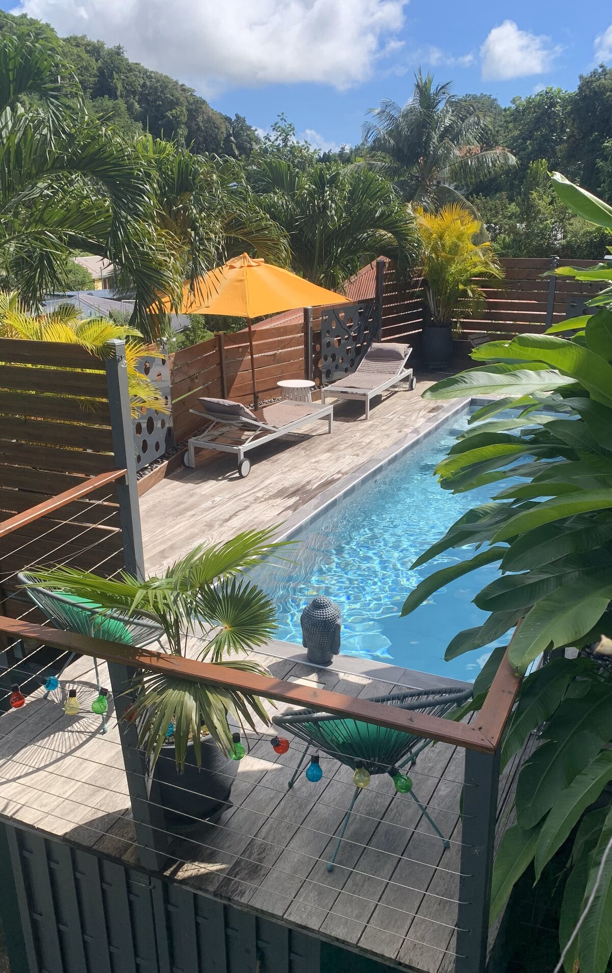 The image displays a serene outdoor pool area surrounded by lush greenery. Sun loungers are positioned beside the pool, accompanied by an orange umbrella providing shade. Wooden decking enhances the relaxing atmosphere, while decorative elements like colorful lights add visual interest.