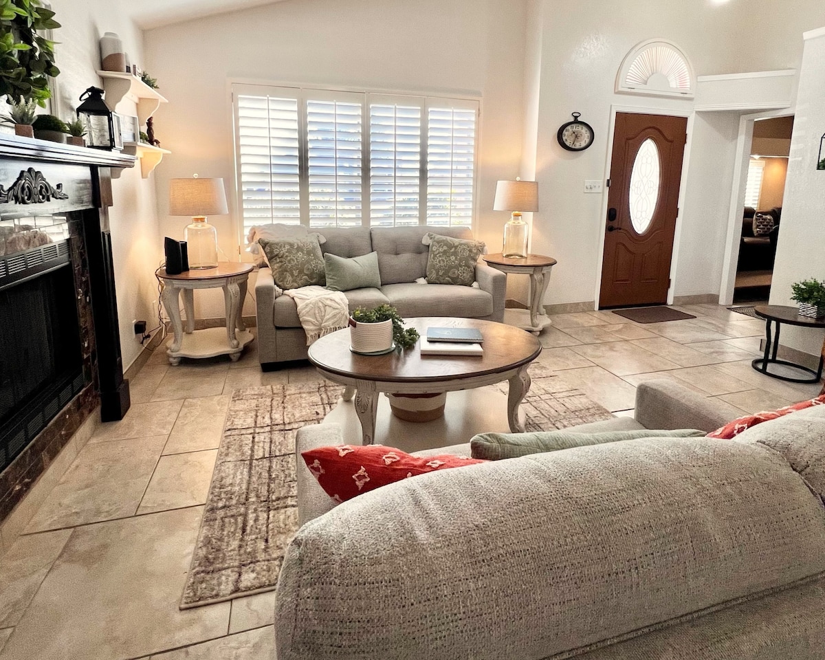 A welcoming living area features a soft gray sofa and a cozy armchair, complemented by decorative pillows. A marble fireplace is flanked by two side tables with lamps. Light filters through plantation shutters, enhancing the inviting atmosphere of the space.