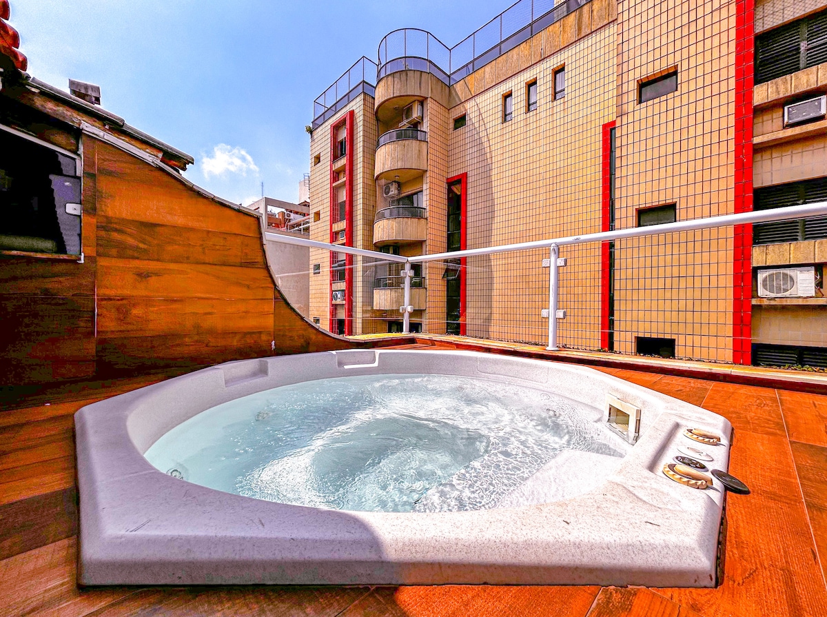 A private jacuzzi situated on a terrace, surrounded by wooden decking. The view includes surrounding buildings, with a clear blue sky above. The jacuzzi features a control panel and is positioned for relaxation in an open-air setting.