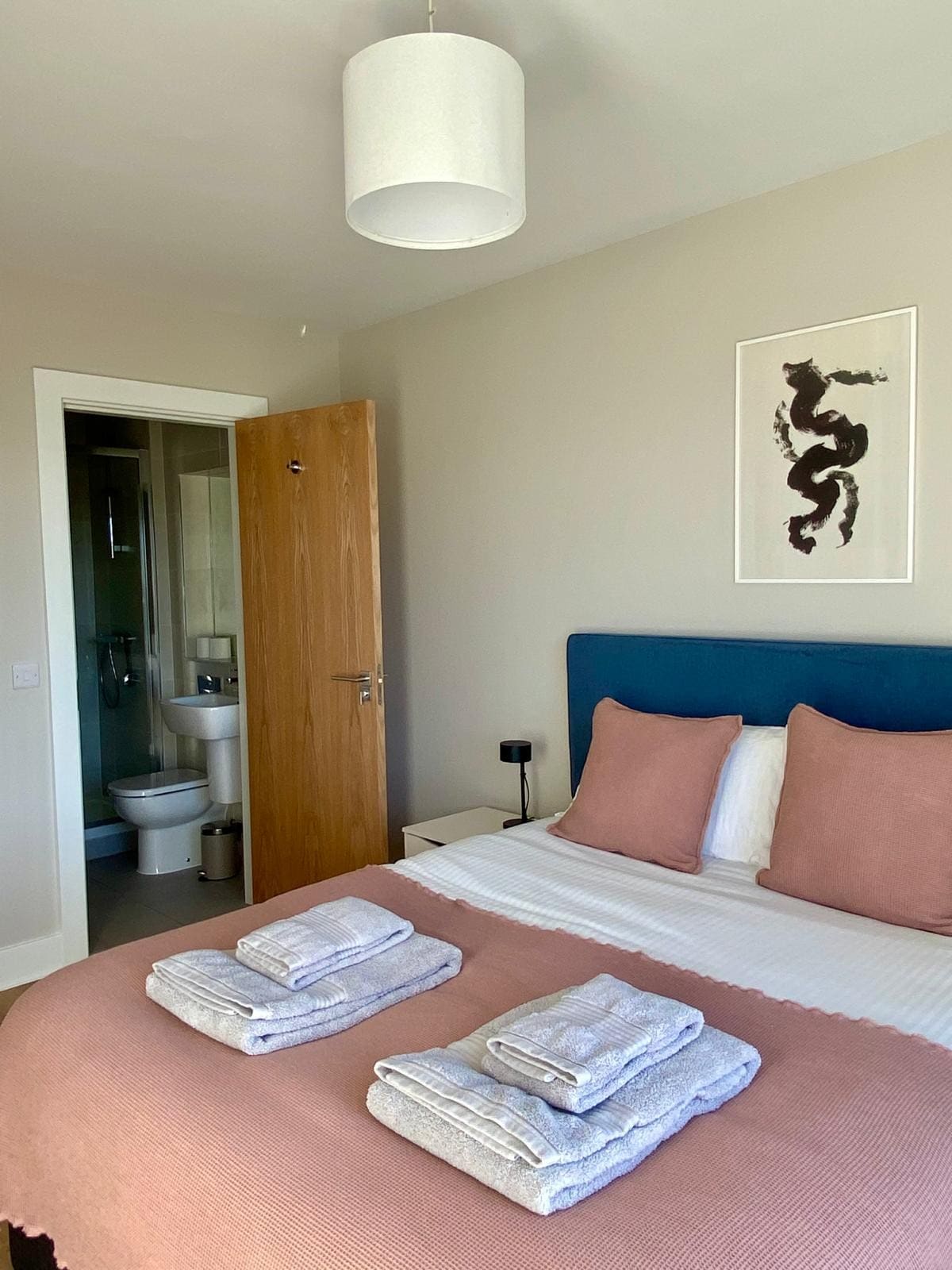 A well-arranged bedroom includes a king-size bed with two neatly folded towels placed on the cover. A framed art piece adorns the wall above the bed. A bathroom is visible through an open door, contributing to an efficient layout.