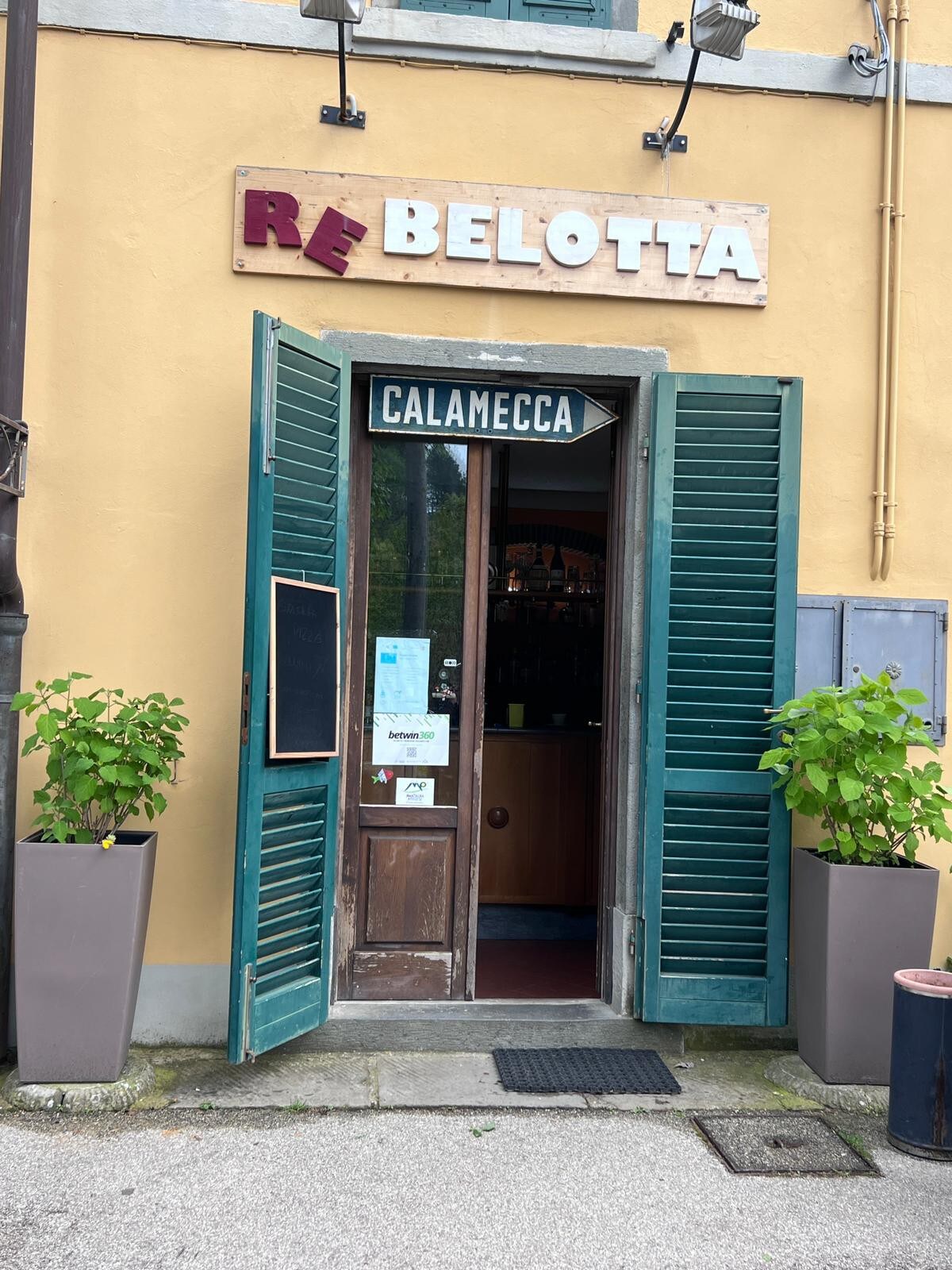 A welcoming entrance is displayed, featuring a wooden door framed by green shutters. The sign above reads 'Re Belotta' with 'Calamecca' below. Potted plants on either side enhance the inviting appearance, while a menu board leans against the wall, visible through the open door.