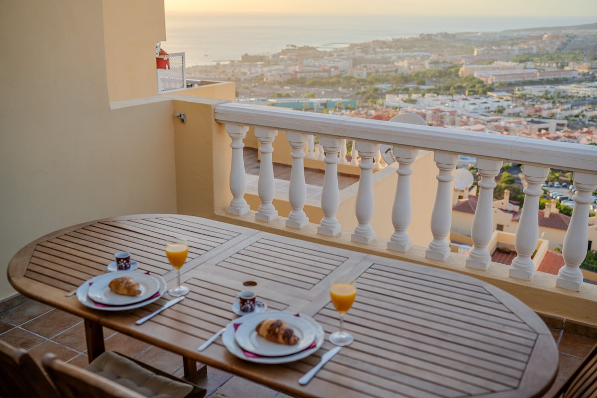 A wooden table is set for two on a terrace, featuring plates with croissants and beverages. The backdrop reveals a panoramic view of the coastline and sunset over the horizon, with various rooftops and greenery visible in the distance.