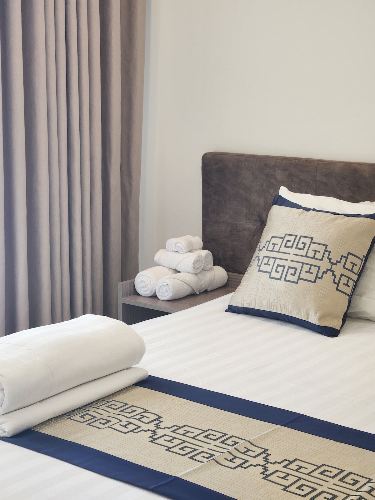 A neatly arranged sleeping area features a spacious bed dressed in neutral tones. A decorative pillow with a geometric pattern rests against the headboard, while a stack of neatly rolled towels is placed on a bedside table. Soft curtains frame the window, allowing natural light to enter.