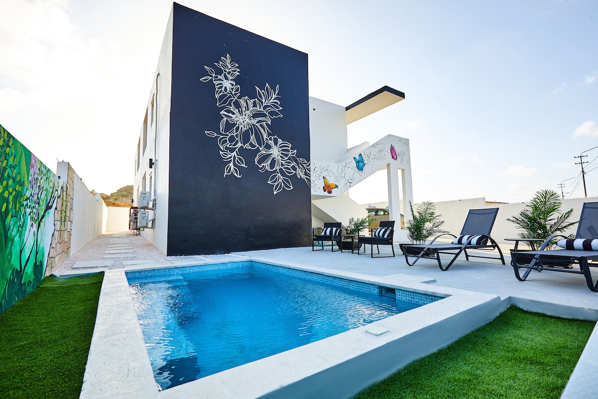A modern building features a wall adorned with artistic murals. A clear blue pool is situated in front, surrounded by lounge chairs on a grassy area, offering a space for relaxation. The bright sky enhances the overall ambiance of the outdoor setting.