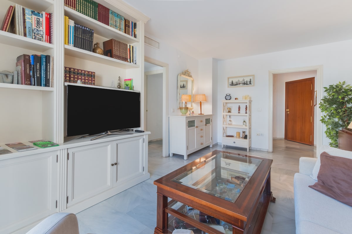 A bright and inviting living area features a modern Smart TV positioned on a white cabinet. A glass coffee table sits in the center, surrounded by a comfortable sofa and shelves lined with books and decorative items. Light-colored walls create a spacious atmosphere.