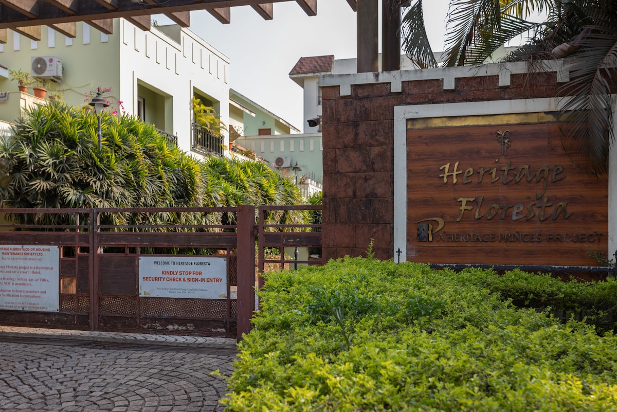 An entrance to a gated community is depicted, featuring a wooden sign that reads 'Heritage Floresta.' Lush green plants and manicured hedges frame the entrance, complemented by a cobblestone pathway leading through the gate. A sense of greenery and privacy is emphasized.