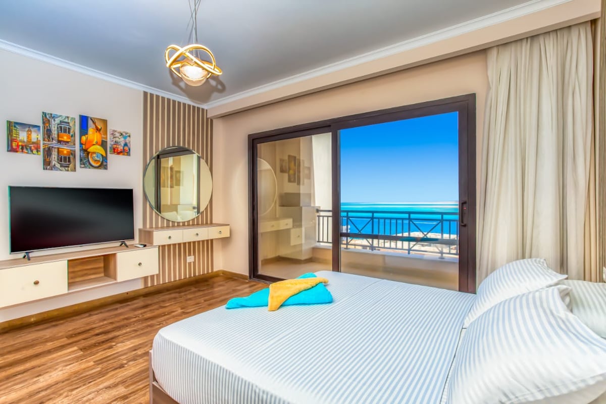 A bedroom features a spacious bed with striped linens, complemented by a large window showcasing a sea view. A modern TV is mounted on the wall, and a circular mirror enhances the room's design. Wooden flooring adds warmth to the space.