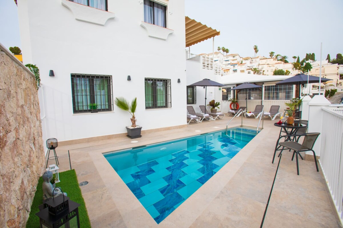 A private outdoor area features a rectangular pool with vibrant blue tiles. Poolside lounge chairs are arranged under large umbrellas, complemented by potted plants. A small table with chairs is positioned nearby, providing a relaxing atmosphere for outdoor enjoyment.