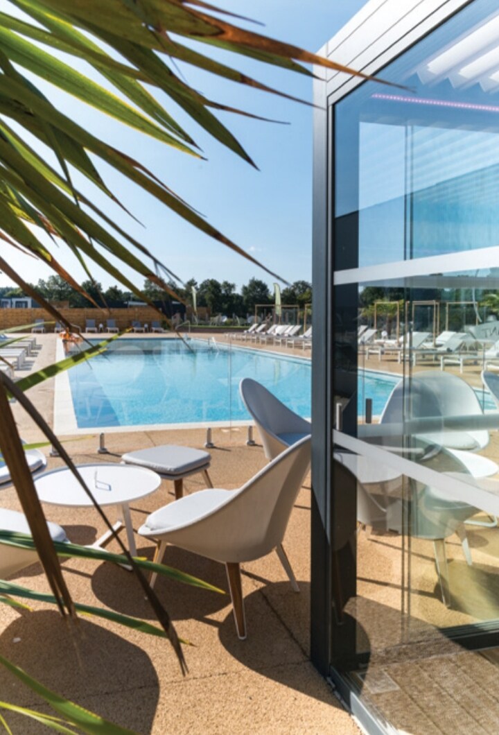 The outdoor pool area is showcased, framed by tropical greenery. Modern lounge chairs are arranged around the sparkling water, with tables positioned nearby for relaxation. Sunlight reflects off the pool surface, creating a serene environment for guests.
