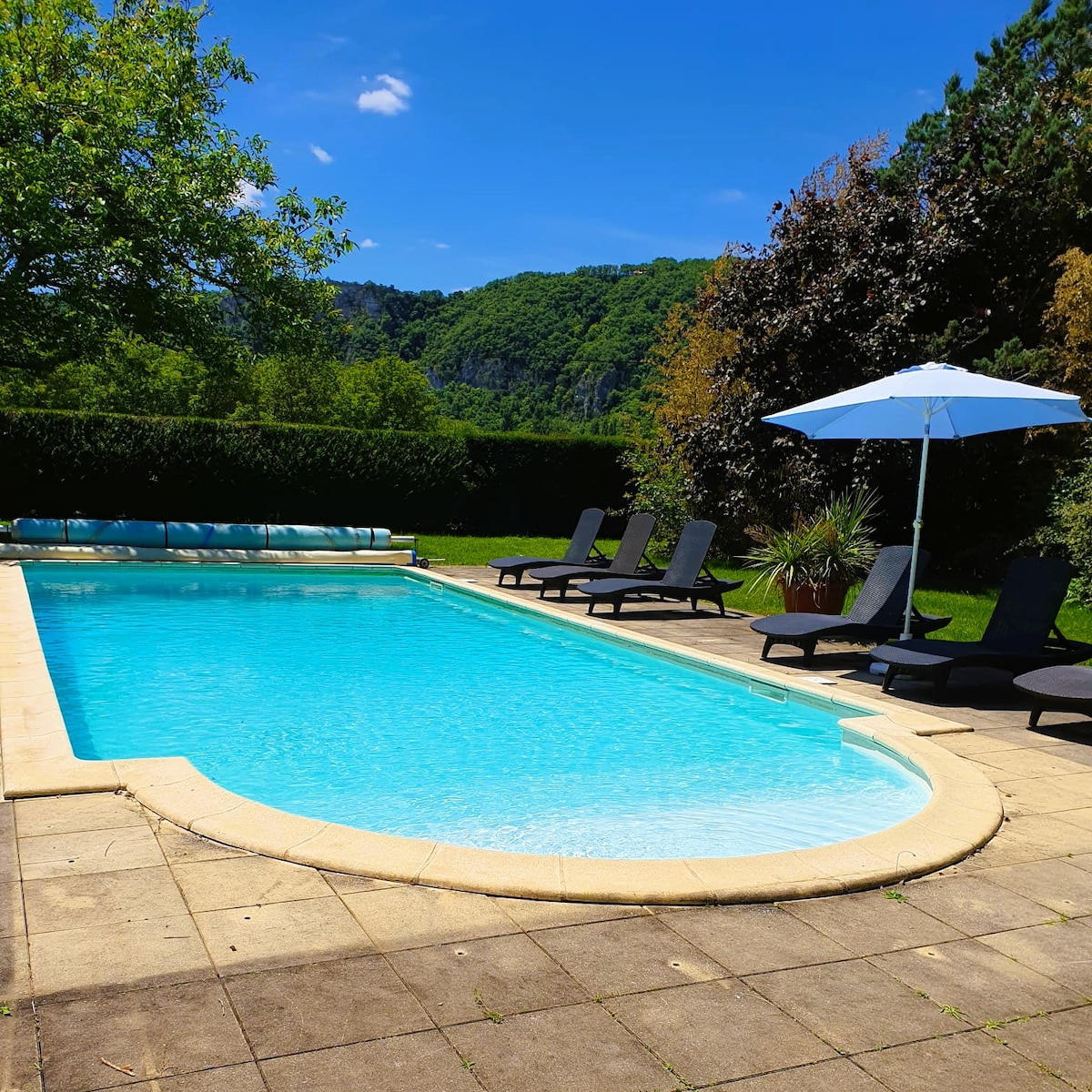A private swimming pool is bordered by a spacious stone patio, featuring several sun loungers positioned for relaxation. A large umbrella provides shade nearby, while lush greenery and rolling hills create a serene backdrop.