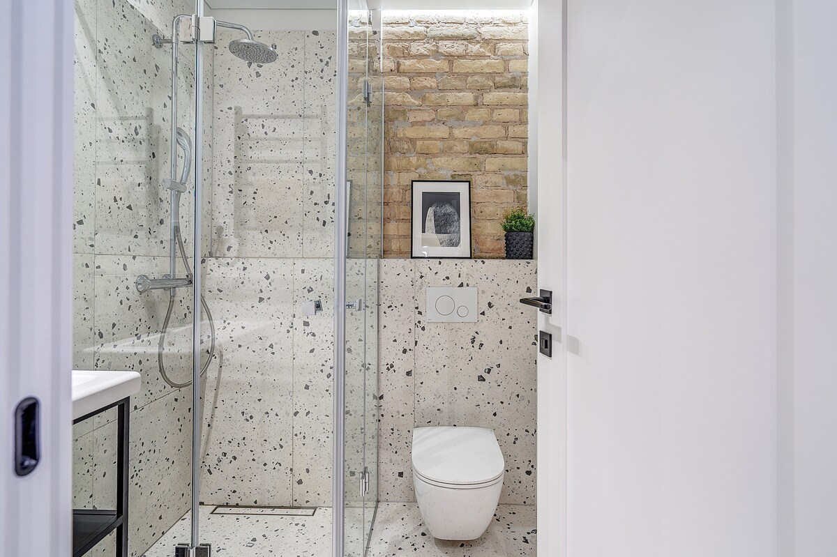 A modern bathroom is observed, featuring a glass shower enclosure and a minimalist toilet. The walls are adorned with speckled tiles and a warm brick accent. A small potted plant and framed artwork provide subtle decor, enhancing the clean and functional design.