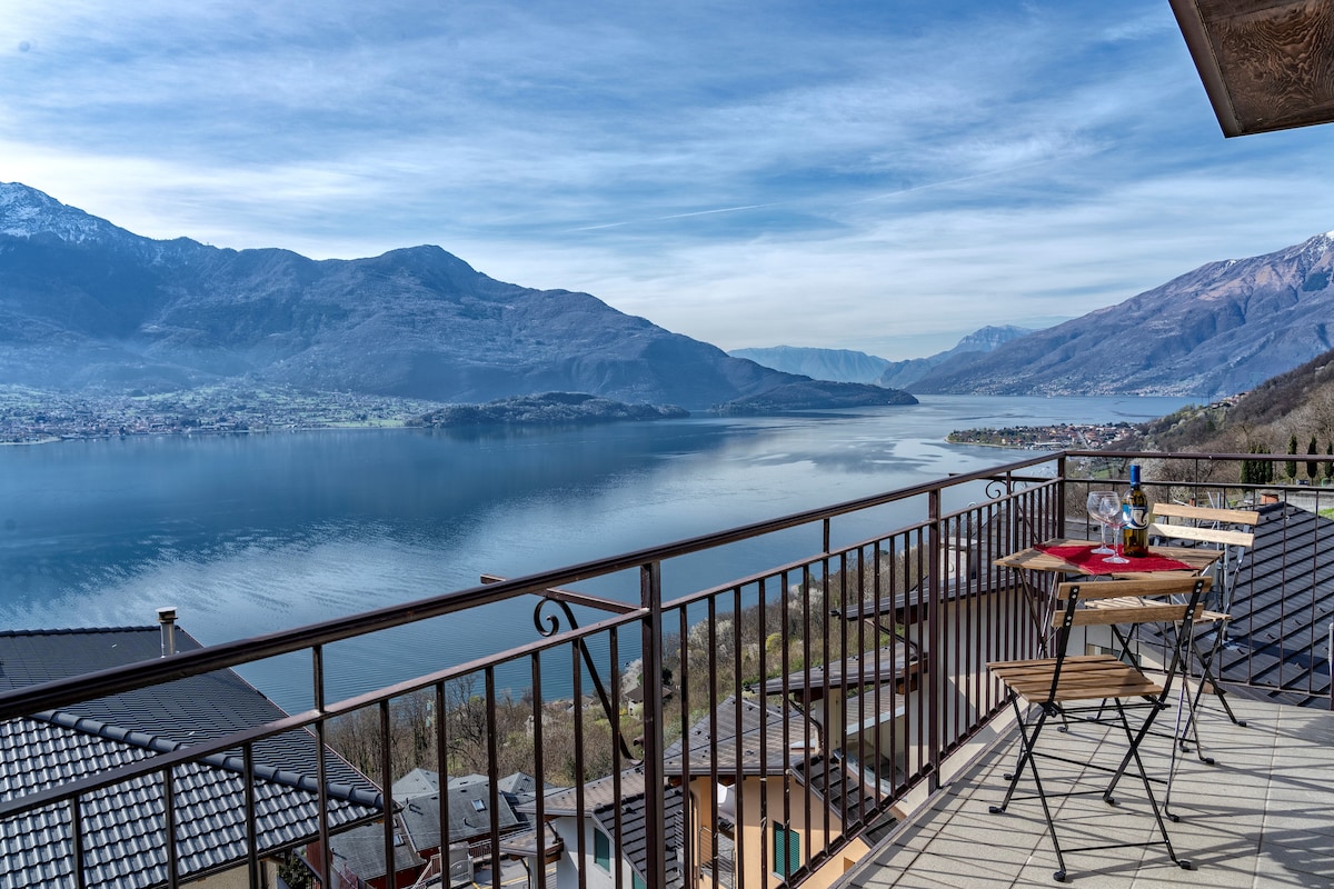 A balcony offers a stunning view of Lake Como and the surrounding mountains. Two wooden chairs and a small table are positioned on the terrace, inviting relaxation while enjoying the serene landscape. The reflection of the mountains on the lake's surface is visible under a clear sky.