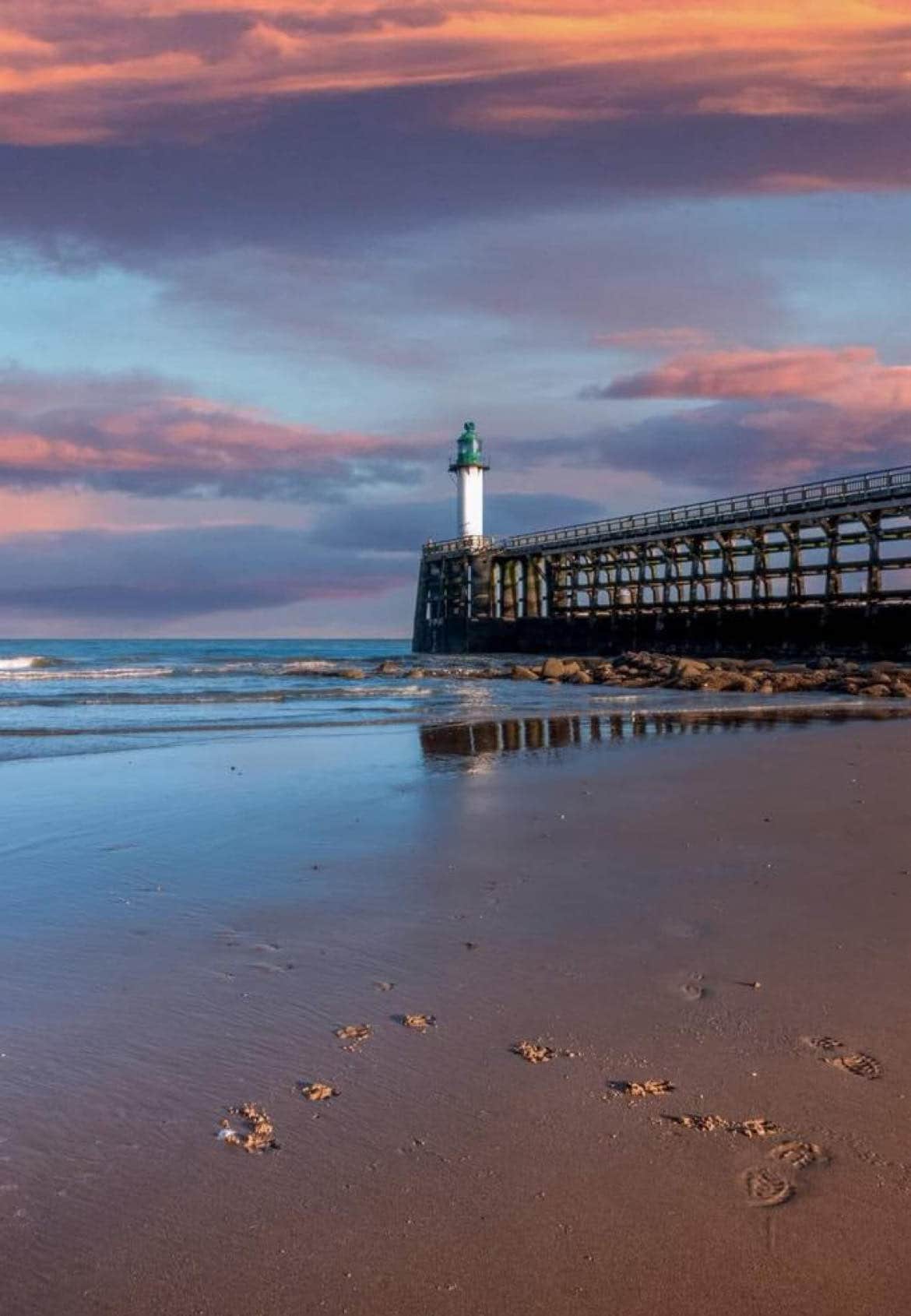 A lighthouse stands on a pier against a vibrant sky at sunset. Soft waves lap at the sandy shore, with footprints leading towards the water. The light beams of the lighthouse reflect on the wet sand, creating a serene coastal scene.
