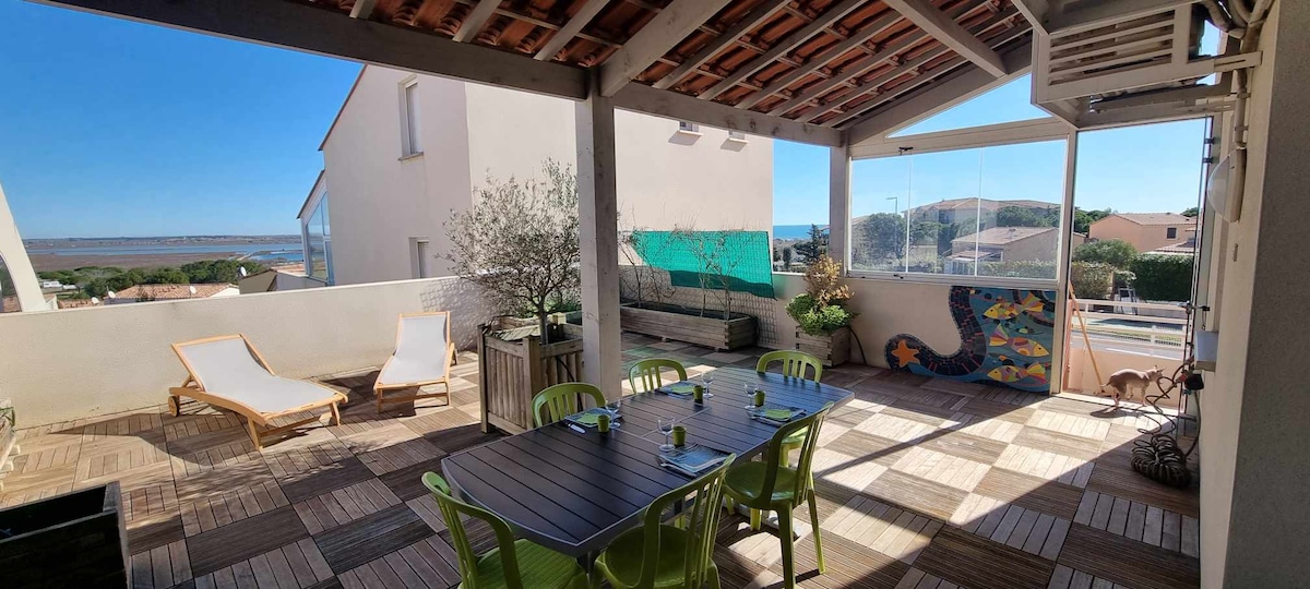 A spacious terrace is presented, featuring a large dining table surrounded by colorful chairs. Two sun loungers are positioned nearby, complemented by a view of the sea in the background. Natural light floods the area, enhancing the terrace's inviting atmosphere.