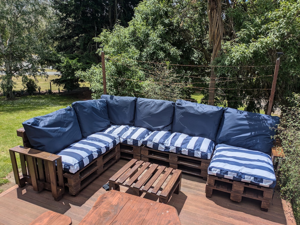 A spacious outdoor lounge area features a sectional sofa made of wooden pallets, adorned with blue and white striped cushions. A wooden coffee table sits in the center, surrounded by lush green plants and trees, creating a comfortable setting for relaxation.