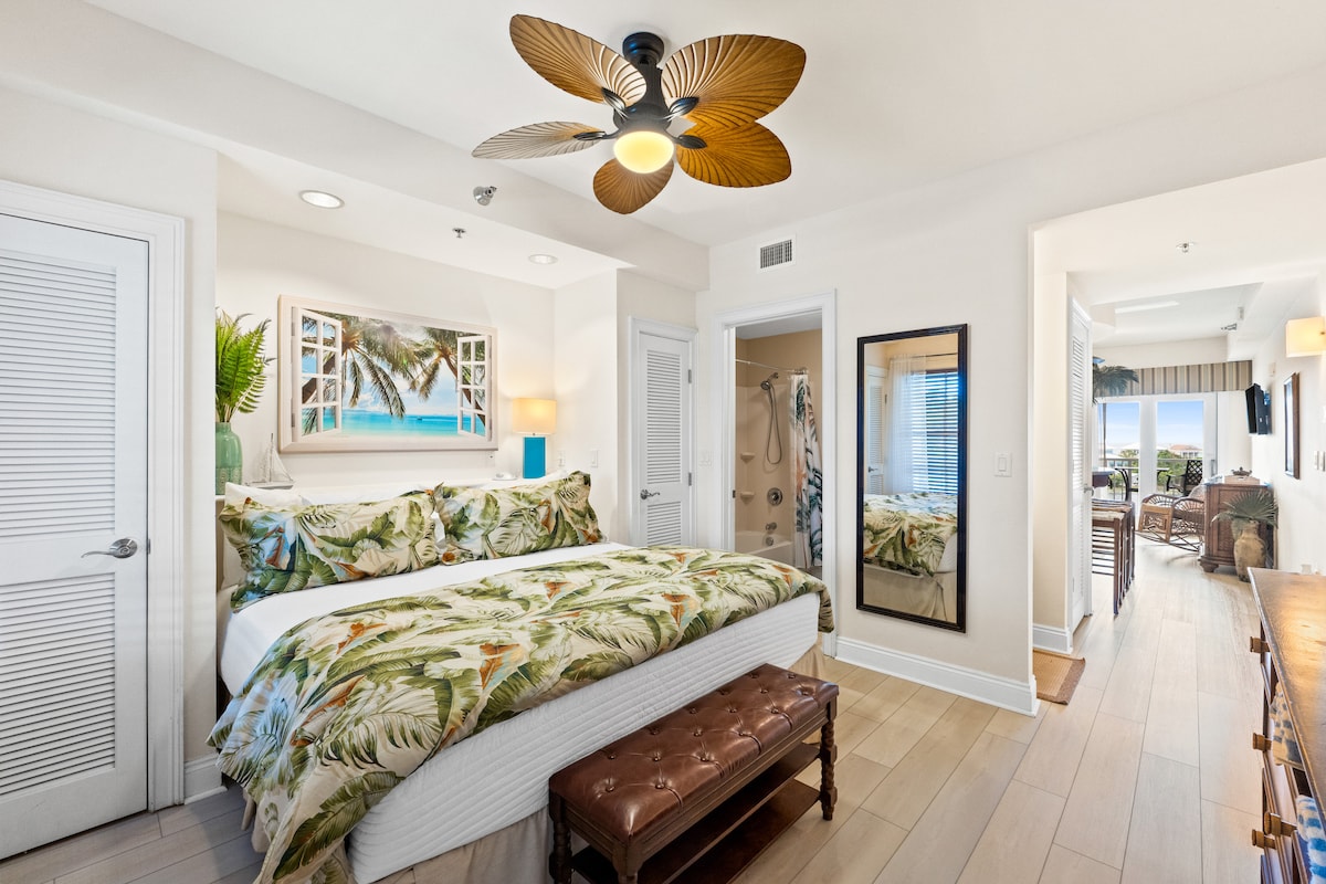 A bright and welcoming primary suite features a spacious bed adorned with tropical bedding. A decorative ceiling fan adds a touch of elegance, while a large mirror reflects the light. The room leads to a hallway, providing glimpses of the nearby bathroom and exterior views.