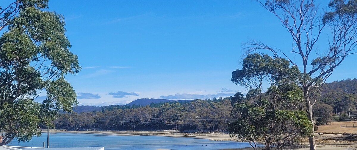 A serene landscape is captured with calm waters reflecting the blue sky. Lush trees frame the scene, and a gentle hillside is visible in the background, evoking a sense of tranquility and connection to nature.