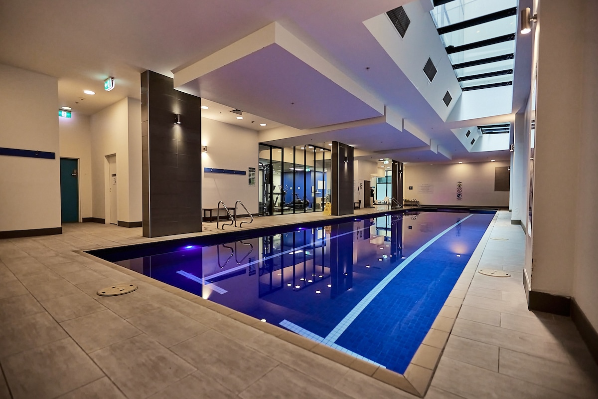An indoor pool area features a sleek, modern design with a long, illuminated swimming pool against a backdrop of large glass windows allowing natural light. Lounge chairs line the edges, creating a relaxing space for guests.