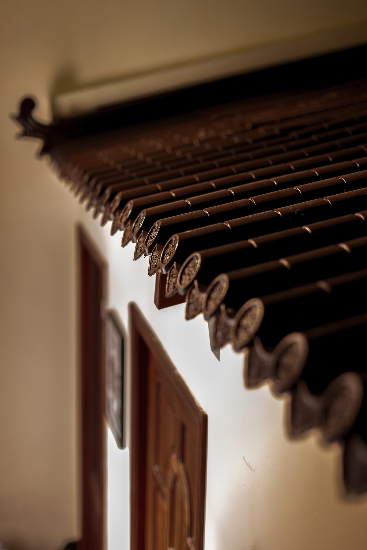 The image captures a close-up view of a traditional tile roof featuring intricate decorative elements. The edges of the tiles are elaborately designed, highlighting the craftsmanship. The muted light enhances the architectural details against the backdrop of the building's white wall and wooden door.
