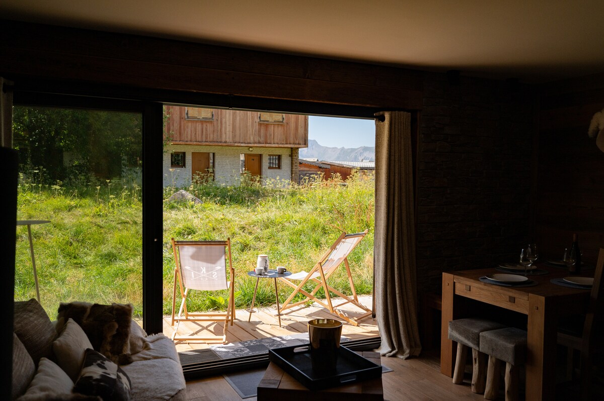 An open space reveals a view of the outdoors through expansive glass doors. Two chairs are positioned on a wooden deck, accompanied by a small table. Lush green grass and a building are visible beyond, creating a serene and inviting environment.