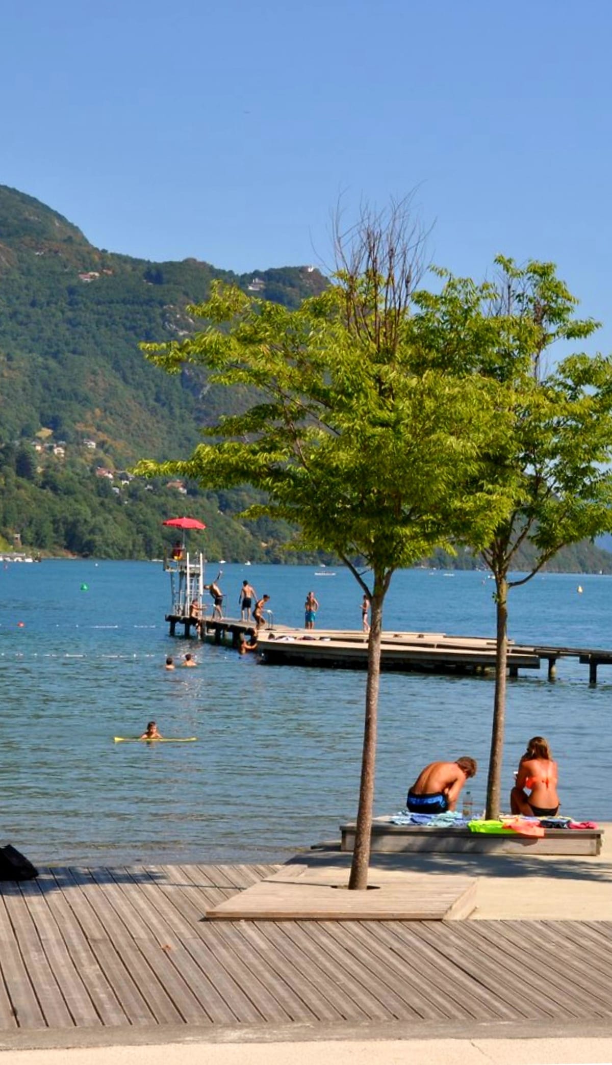 The image captures a serene lake scene with a wooden pier extending into calm water. Nearby, two trees provide shade, while individuals swim and relax in the water. Mountains rise in the background, complemented by a clear blue sky.