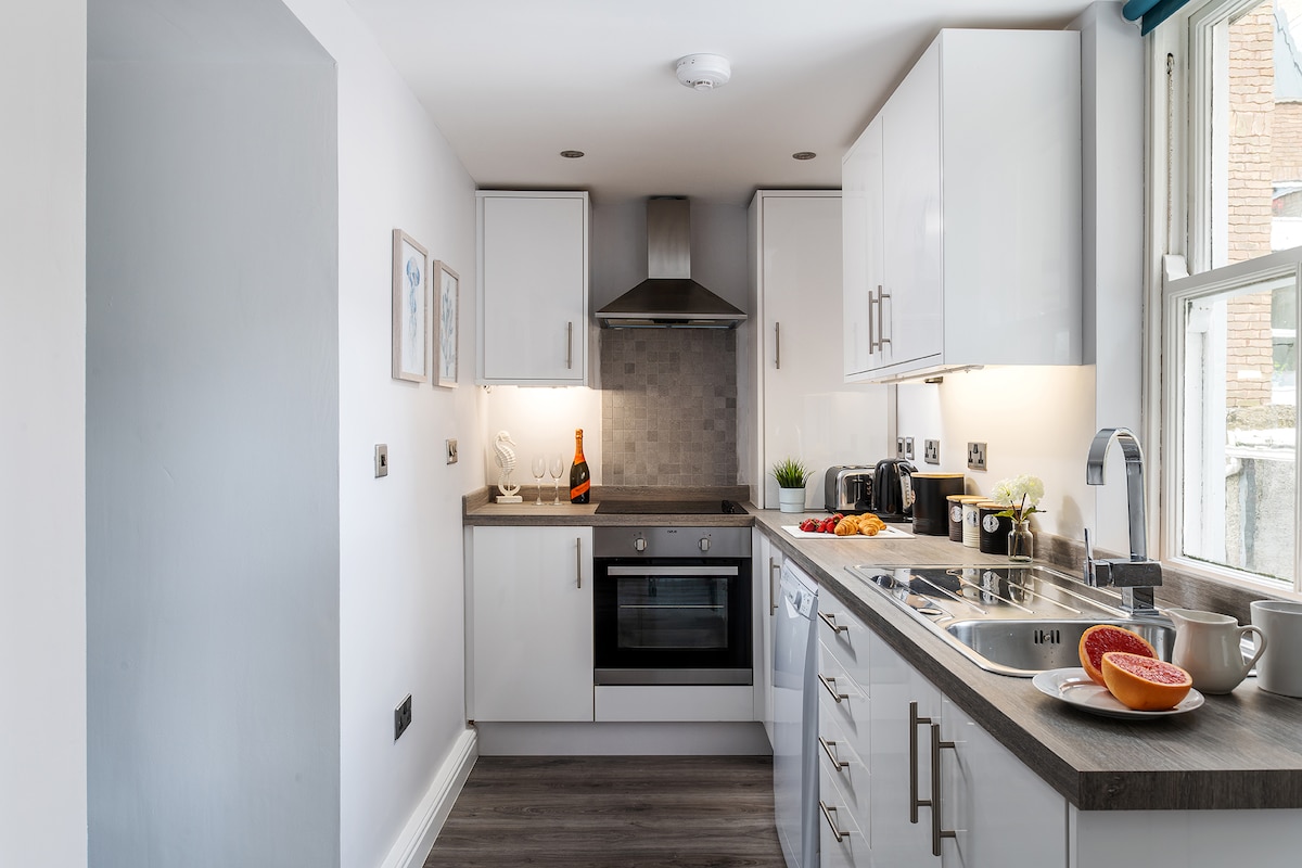 A modern kitchen equipped with essential appliances, including an oven, microwave, and toaster. Light-colored cabinets contrast with the dark countertop. A grapefruit sits on a plate near the sink, adding a touch of color. Natural light streams in through a large window.