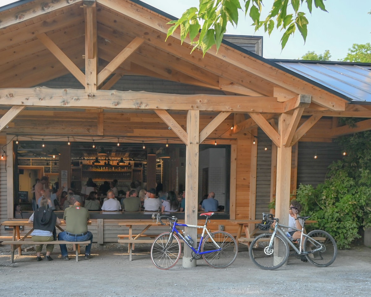 A rustic wooden structure with a large roof overhang is depicted, featuring a spacious outdoor seating area. Long picnic tables line the porch, while several bicycles are parked nearby. Strings of lights are hung above, adding warmth to the inviting environment.