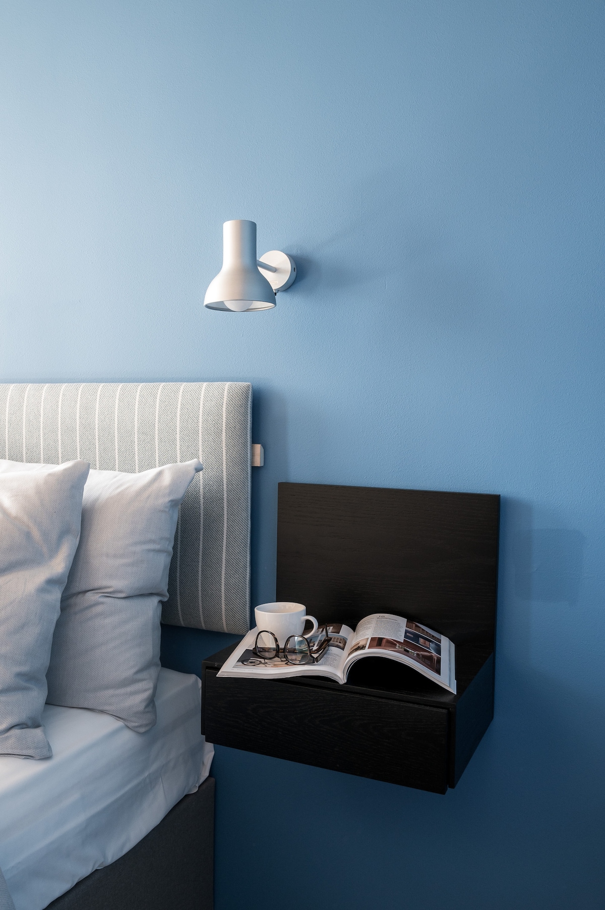 A bedside area features a soft gray headboard and a dark nightstand. A book is open, accompanied by a pair of glasses and a coffee cup. The wall has a modern lamp, casting light onto the calming blue background.