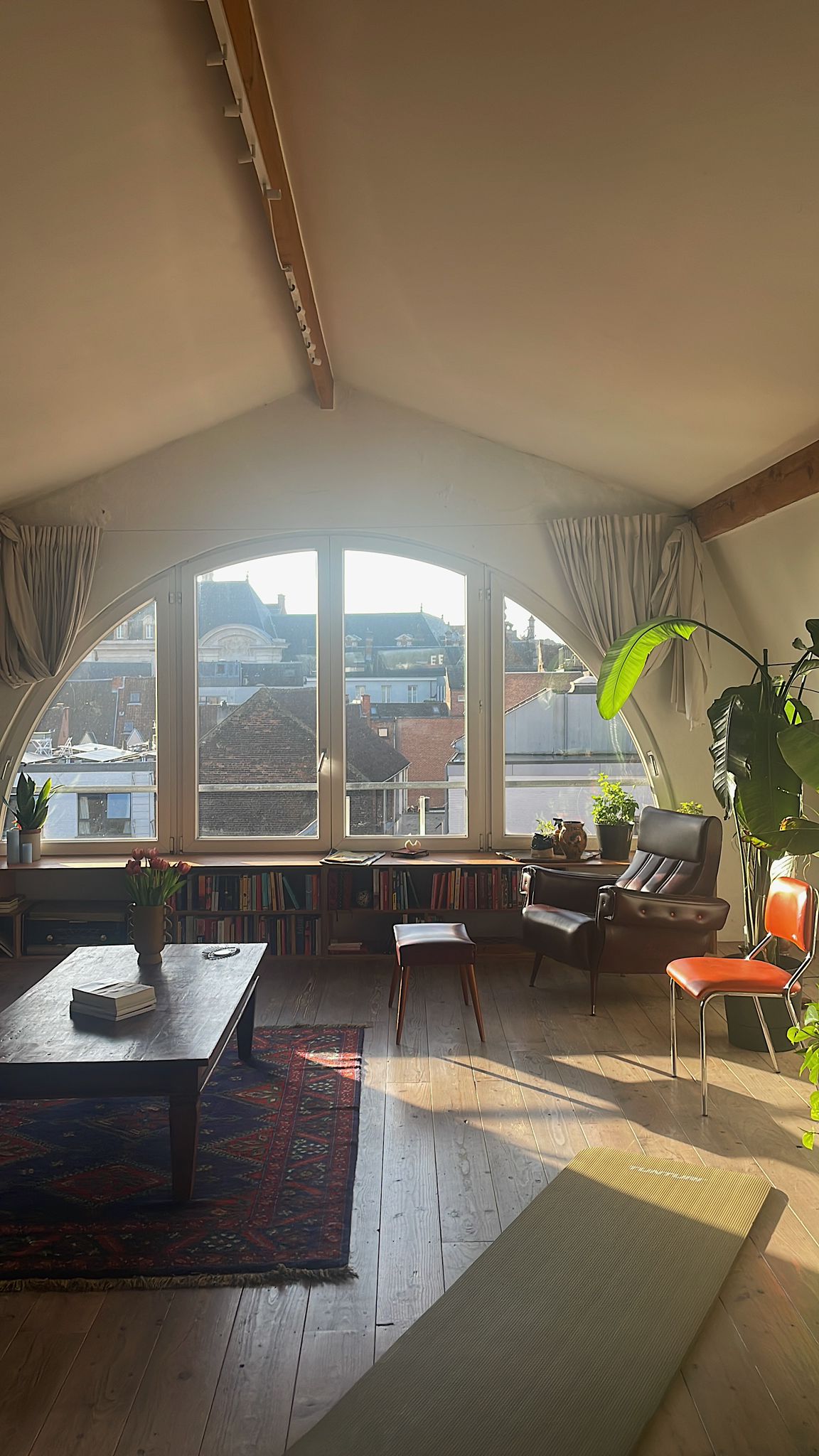 A bright living area features large, arched windows that offer views of the cityscape. A cozy seating arrangement includes a dark armchair and a small wooden chair, complemented by a wooden coffee table. Shelves filled with books line one wall, and plants add greenery to the space.