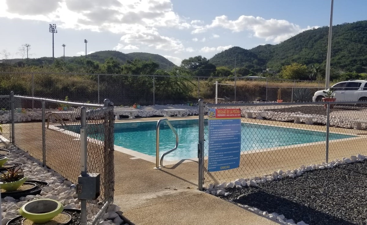 A fenced pool area is featured with clear water and a set of steps leading into the water. Surrounding the pool are landscaped areas with rocks and potted plants, while a backdrop of rolling hills and scattered clouds is visible.