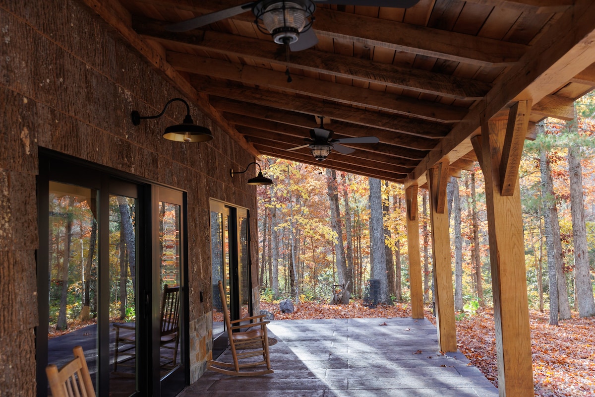 A spacious covered porch features rustic wooden rocking chairs and overhead ceiling fans. The large glass doors reflect the forested surroundings, where autumn leaves create a colorful carpet on the ground, conveying a sense of tranquility and connection to nature.