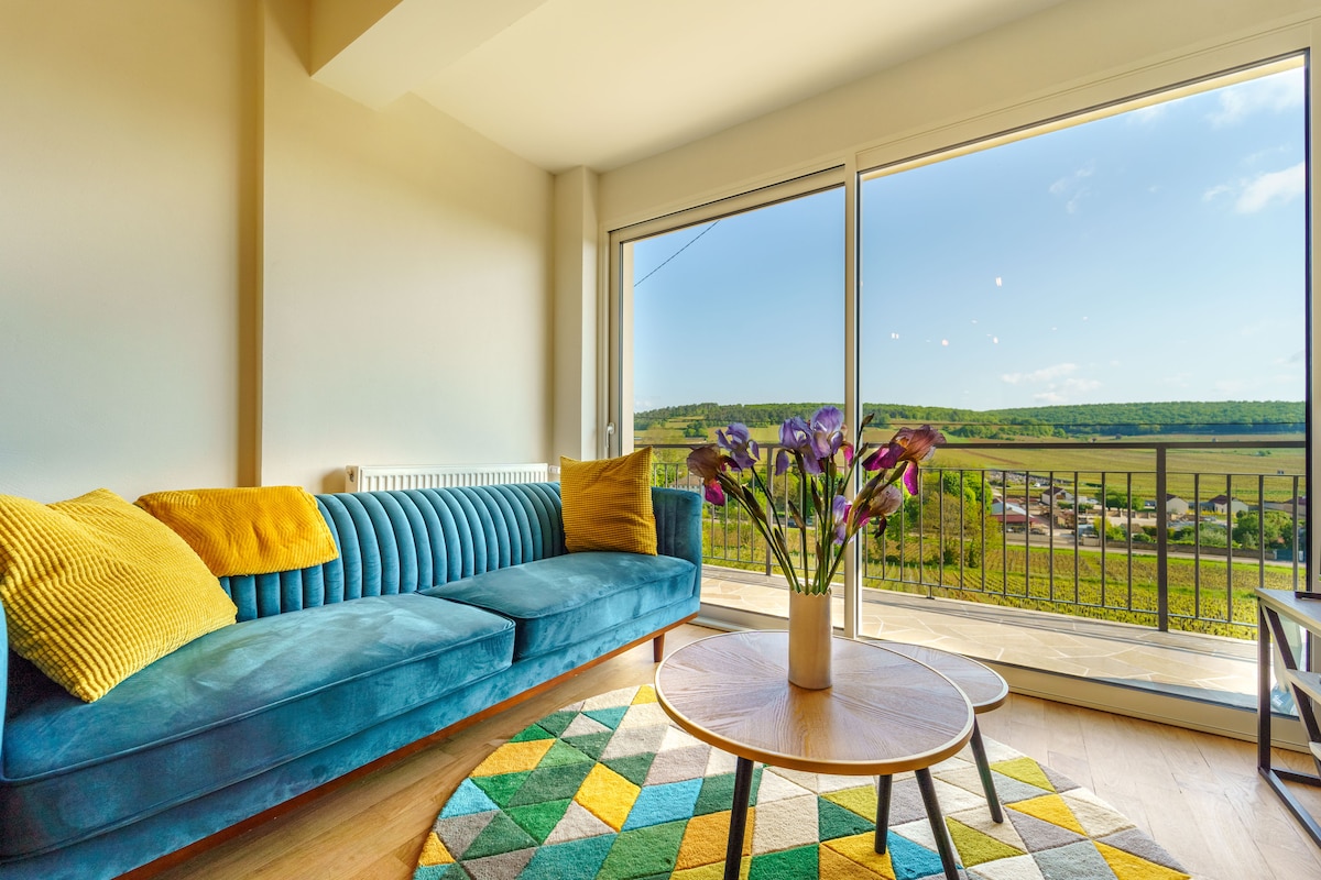 A bright living space features a blue couch adorned with yellow cushions, complemented by a geometric patterned rug. A coffee table sits in front, and large windows provide views of the surrounding vineyards and countryside, allowing natural light to illuminate the room.