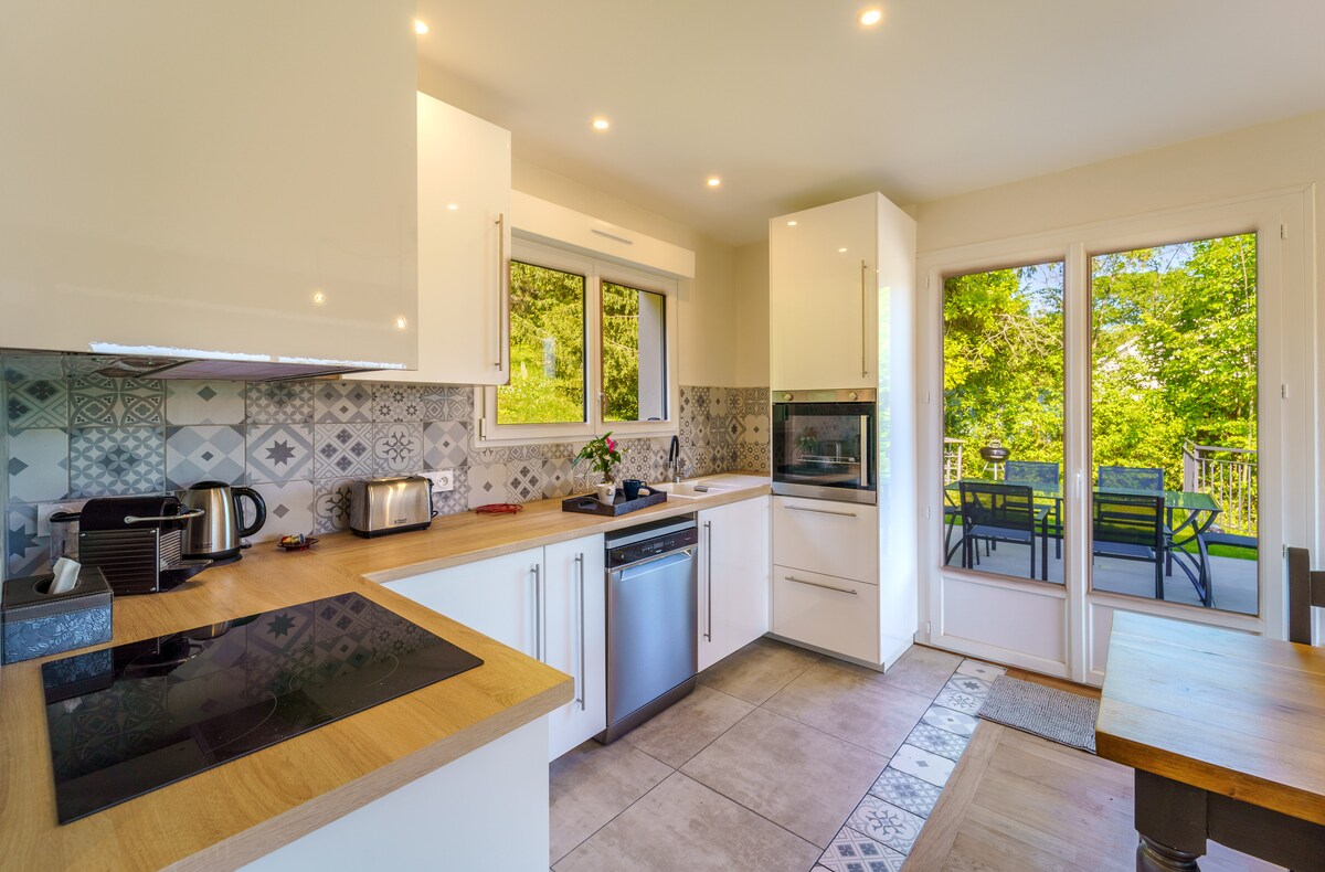 A modern kitchen features sleek cabinetry and a large window overlooking greenery. Stainless steel appliances, including a dishwasher and microwave, are visible. The kitchen is complemented by a wooden countertop and patterned backsplash, with a dining area nearby for shared meals.