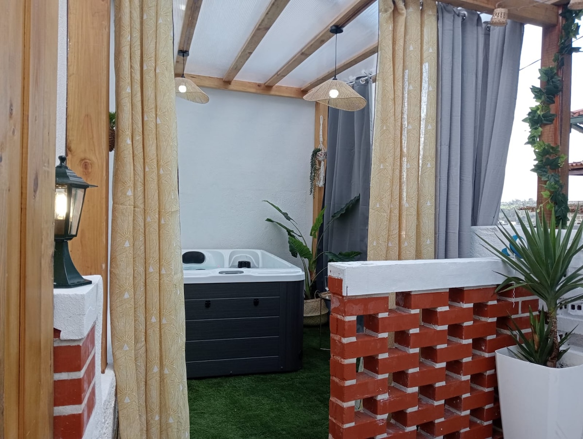 A private jacuzzi is positioned in a cozy open space, surrounded by wooden beams and soft canopy curtains. The area features green artificial grass and potted plants, while warm lighting fixtures provide a gentle glow.