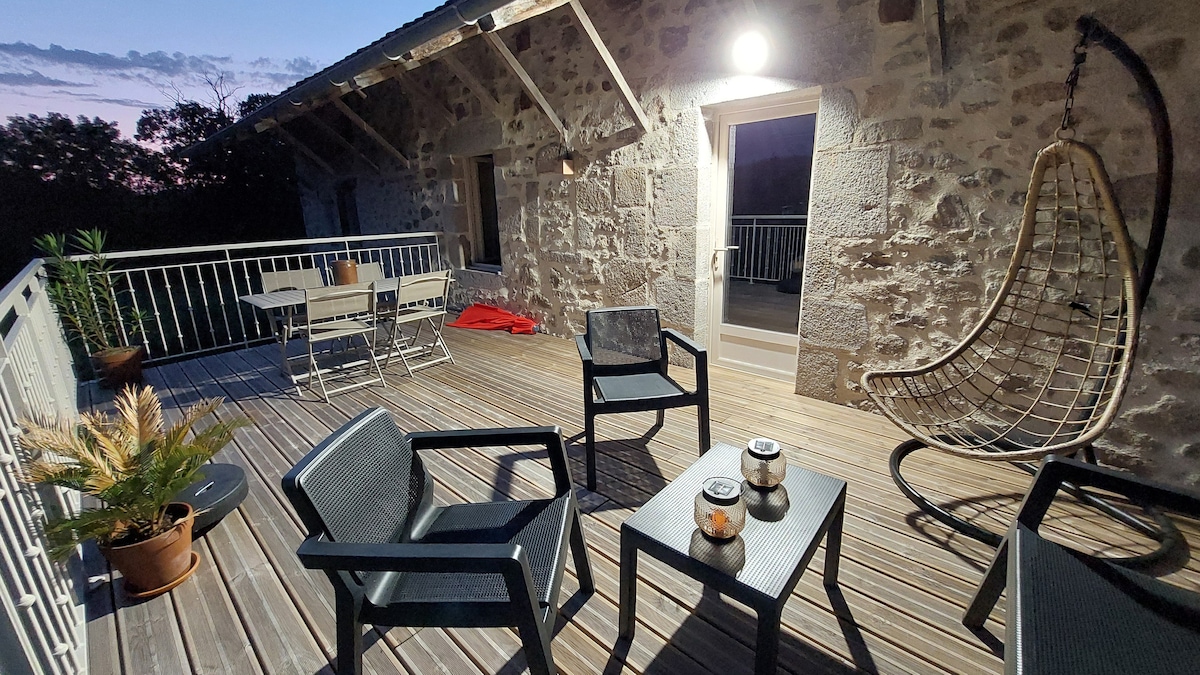 A spacious terrace is presented with wooden flooring and stone walls. Outdoor seating is arranged, featuring a hanging chair and two armchairs beside a table with decorative elements. Soft lighting enhances the cozy evening ambiance, while the surrounding nature can be glimpsed in the background.